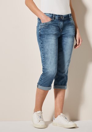 Pantalones capri de mezclilla azul claro con un acabado desteñido, con puños enrollados y dos bolsillos laterales, combinados con zapatillas blancas.