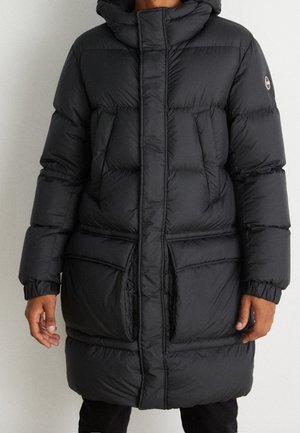 Musta pitkä toppatakki hupulla, vetoketjulla edessä, läppätaskuilla ja joustavilla hihansuiden resoreilla, jota käyttää henkilö, jonka kädet näkyvät.