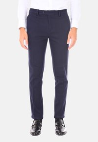 Pantaloni slim-fit blu navy con parte frontale liscia, passanti per cintura e chiusura con un solo bottone, abbinati a scarpe in pelle lucida nere.