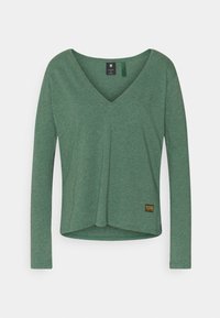 Grünes Langarmshirt mit V-Ausschnitt, aus weichem, strukturiertem Stoff, mit leicht ausgestelltem Saum und einem kleinen Logo-Patch am unteren Rand.