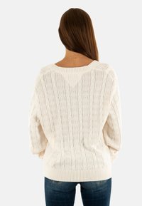 Donna con lunghi capelli castani, che indossa un maglione bianco a treccia e jeans blu, girata di spalle contro uno sfondo neutro.