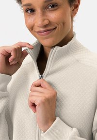 Veste zippée beige clair avec un motif matelassé texturé, col haut et poignets côtelés. Le mannequin dézippe la veste, mettant en valeur le design.