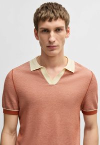 Ljus terrakotta kortärmad polo t-shirt med krämfärgad V-ringad krage och kontrasterande röda ärmslut, med en texturerad stickad tyg.