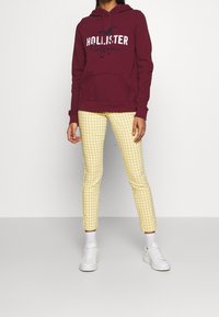 Felpa con cappuccio color mogano con logo, tasca frontale e coulisse. Pantaloni gialli a quadretti. Scarpe da ginnastica bianche con suole imbottite e senza marchi visibili.