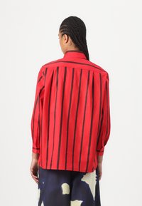 Camicia rossa con strisce verticali nere, polsi abbottonati e colletto, con una vestibilità rilassata e pieghe sul retro per un tocco di trama in più.