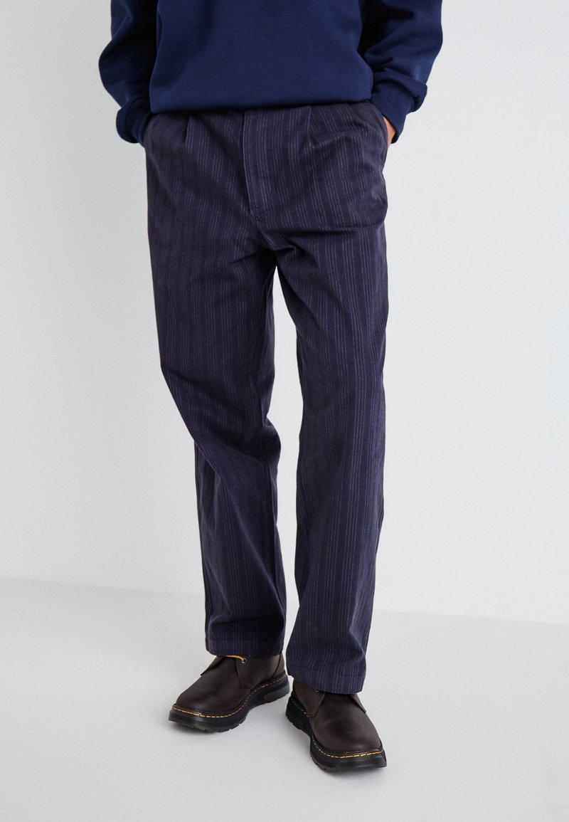 Navy corduroy trousers with vertical pinstripes, featuring a relaxed fit and pockets. Paired with dark brown leather shoes.