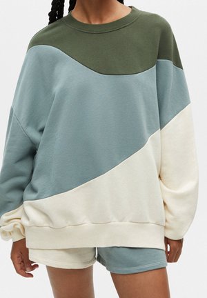 Person iført oversized color-block sweatshirt i olivengrøn, lyseblå og off-white med matchende shorts.