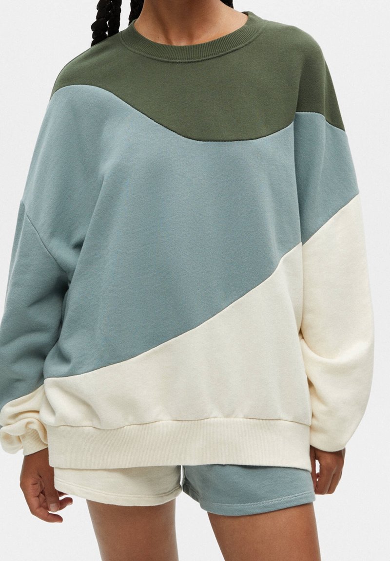 Personne portant un sweat-shirt oversize à blocs de couleurs en vert olive, bleu clair et blanc cassé avec un short assorti.