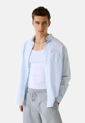Giovane uomo con indosso una camicia celeste a bottoni sopra una canottiera bianca e pantaloni da tuta grigi con coulisse, di fronte, su sfondo bianco.
