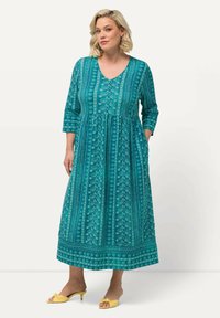 Vestido maxi en color verde azulado con escote en V, mangas de tres cuartos y estampado floral. Presenta una cintura fruncida y bolsillos laterales, combinado con zapatos amarillos.