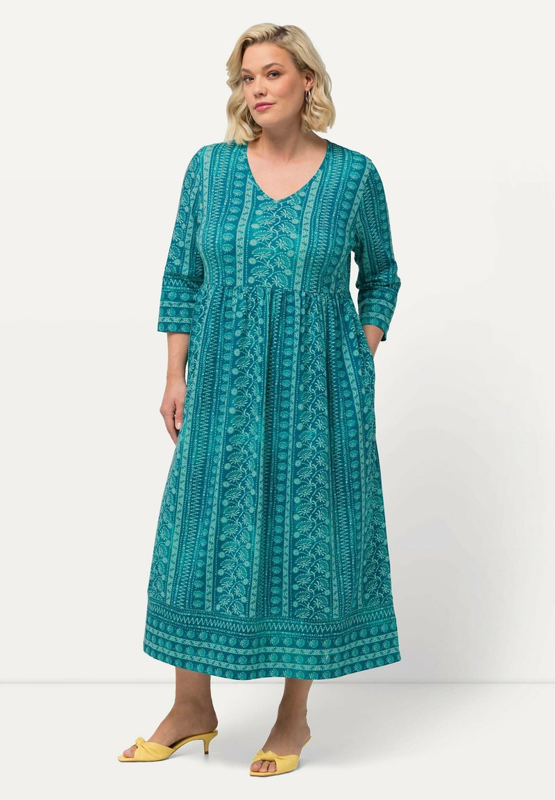 Vestido maxi en color verde azulado con escote en V, mangas de tres cuartos y estampado floral. Presenta una cintura fruncida y bolsillos laterales, combinado con zapatos amarillos.