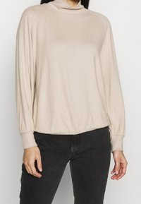 Haut beige à manches longues avec col montant, en tissu doux. Présente une coupe décontractée et des poignets côtelés, avec un ourlet froncé.