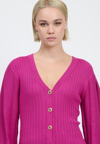 Fuchsia strikket cardigan med dyb V-udskæring, ribbet tekstur og tre guldknapper foran, med blonderdetaljer på ærmerne.