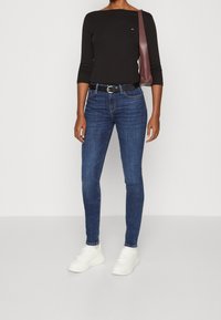 Top oscuro de manga larga combinado con jeans ajustados azules y zapatillas blancas. El conjunto incluye un cinturón negro y un bolso de hombro burdeos.