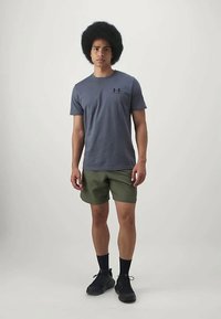 Jauns vīrietis stāv, ģērbies pelēkā Under Armour t-kreklā, olīvu zaļa šortos, melnās zeķēs un melnās kedās pret vienkāršu fona.