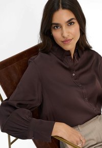 Blusa di raso marrone con colletto classico, chiusura con bottoni e maniche plissettate. Texture liscia, abbinata a pantaloni beige e una sedia marrone.