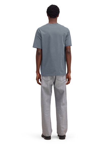 Personne debout de dos, portant un t-shirt bleu ardoise uni, un jean gris clair à jambes larges et des chaussures noires sur un fond blanc.
