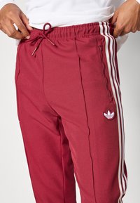 Sarkani sporta bikses ar baltām un zelta svītrām, ar elastīgu jostasvietu ar auklu un mazu izšūtu logo sānos.