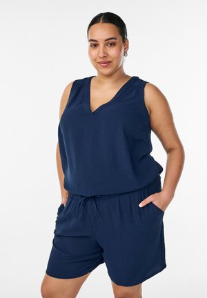 Donna con capelli scuri raccolti in una coda di cavallo, indossa un top blu navy senza maniche con scollo a V e pantaloncini coordinati, mani in tasca, in piedi su sfondo bianco.