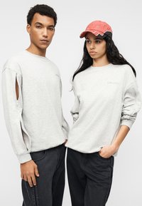 Γκρι φούτερ με φερμουάρ στο πλάι, σε συνδυασμό με μαύρο παντελόνι. Ένα μοντέλο φοράει κόκκινο καπέλο με λευκές λεπτομέρειες.