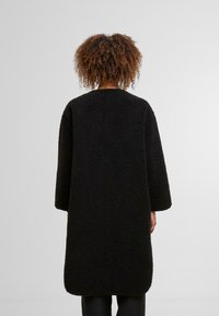 Cappotto nero, oversize, realizzato in un materiale texturizzato e peloso, con maniche lunghe e scollo rotondo, che arriva fino a metà coscia con un orlo dritto.