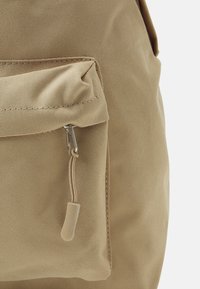 Mochila de lona beige con un acabado texturizado, que cuenta con un bolsillo delantero con cremallera y un acento en el tirador de la cremallera.