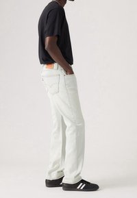 Mężczyzna w jasnych dżinsach, czarnej koszulce, czarnych trampkach z białymi paskami, z rękami w kieszeniach, stojący w profilu na jednolitym tle.