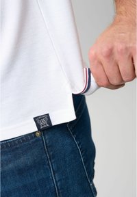 Hvid polo-shirt med kontrasterende røde og blå striber på kraven og et logomærke ved kanten. Båret over mørke denim jeans.