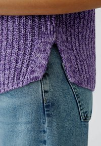 Lila gestrickter Pullover mit gerippter Textur, ausgestattet mit einem Seitenschlitz, kombiniert mit hellblauen Jeans. Fokus auf die Stoffdetails und den Farbharmonien.