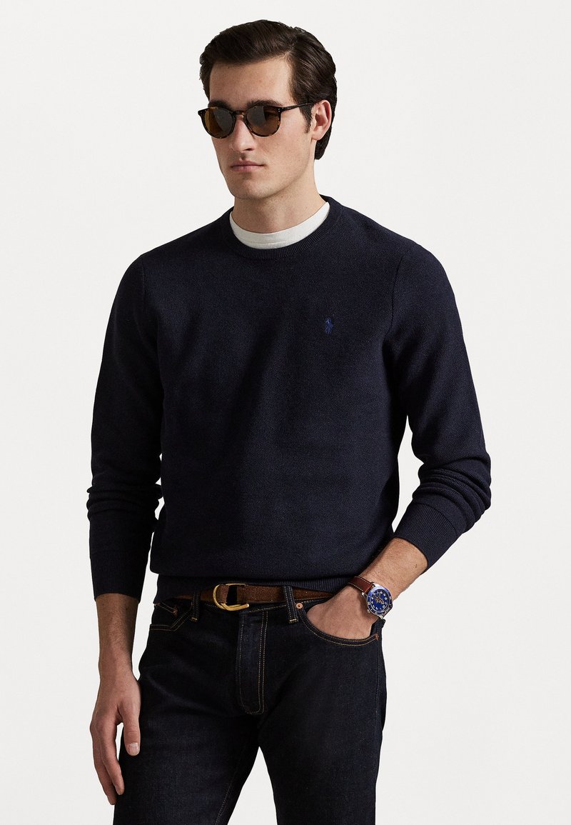 Suéter náutico de cuello redondo hecho de tejido de punto texturizado, combinado con jeans oscuros. Los accesorios incluyen un cinturón y un reloj marrones, complementados con gafas de sol.