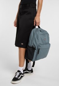 Mochila de color azul claro con un exterior texturizado, compartimentos con cremallera y detalles de malla negra, combinada con pantalones cortos negros y zapatillas deportivas.