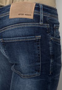 Mörkblå denimjeans med en texturerad brun läderlapp märkt "ANTONY MORATO" på baksidan; har en klassisk femficksdesign och sömnadsdetaljer.