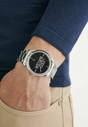 Montre en acier inoxydable argenté avec un cadran noir comportant du texte blanc, portée au poignet d'une chemise à manches longues bleu marine.