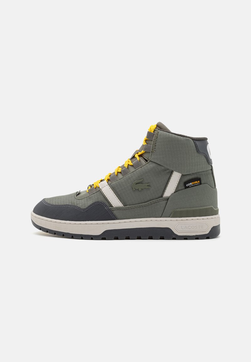 Zapatillas altas de tela verde oliva con cordones amarillos, que presentan detalles en gris, suela texturizada y la marca Lacoste en el costado.