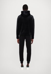 Felpa con cappuccio nera realizzata in materiale morbido, abbinata a pantaloni della tuta neri e scarpe bianche con accenti verdi. Texture liscia, vestibilità rilassata.