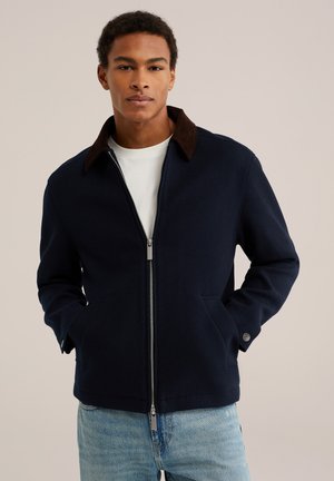 Jeune homme portant une veste zippée bleu marine avec un col marron foncé, une chemise blanche en dessous et un jean bleu clair, les mains dans les poches de la veste.