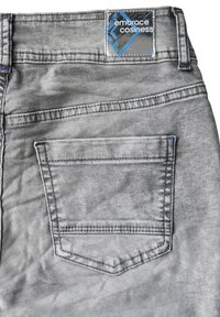 Jeans en denim gris avec une petite poche arrière, des coutures contrastées et une étiquette de marque rectangulaire en bleu et noir. Surface de tissu texturée.
