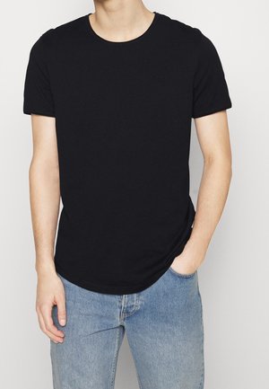 Homme portant un t-shirt noir uni à manches courtes et col rond, et un jean en denim bleu clair avec une main dans la poche, sur fond blanc.