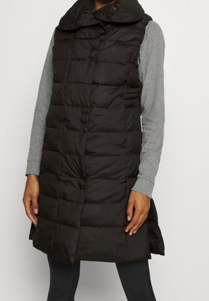 Femme portant une doudoune sans manches noire matelassée par-dessus un t-shirt gris à manches longues, avec un pantalon noir sur un fond uni.