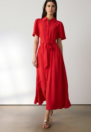 Frau steht vor einer schlichten Wand und trägt ein rotes Midi-Kleid mit Knopfleiste, kurzen Ärmeln, gebundener Taille und beigen Riemchensandalen.