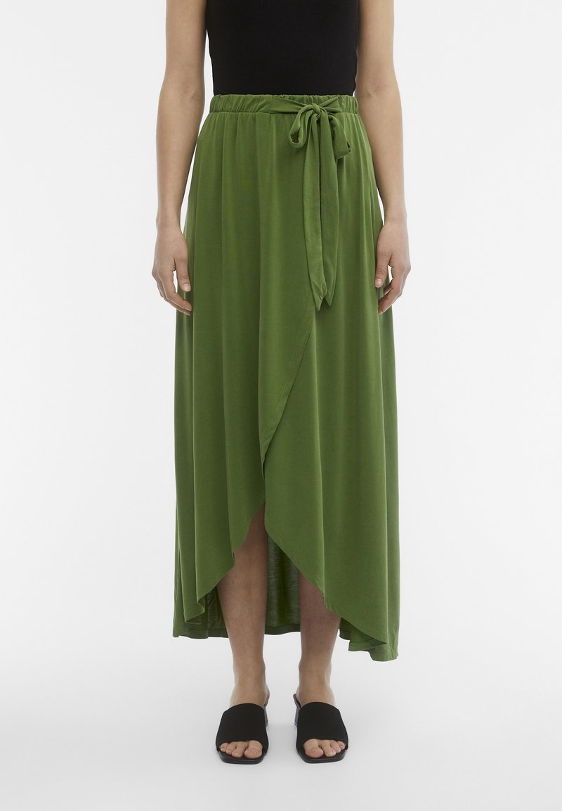 Femme portant une jupe portefeuille verte taille haute nouée à la taille, associée à des sandales mules noires ouvertes et un haut noir sans manches.