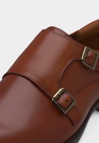 Chaussure à boucle monastique en cuir marron avec deux boucles dorées et coutures détaillées sur fond blanc.