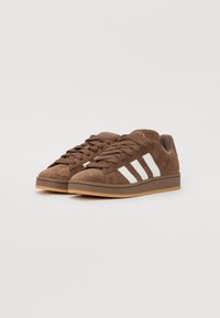 Brown suede sneakers with white three-stripe design, round toe, and brown rubber sole. Features flat laces and padded collar.