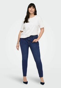 Bílý tričko s krátkým rukávem a krajkovým lemem spárované s tmavě modrými skinny džínami. Modelka stojí s důvěrou, s jednou rukou na bocích.