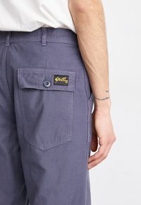 Gray cotton pants with a side pocket, featuring a button closure and a black label with yellow text. Smooth texture and tailored fit.