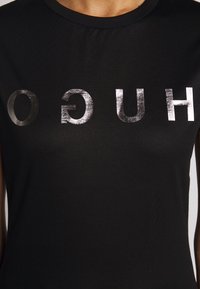 Camiseta de algodón negra con letras impresas en plata metálica que deletrean "HUGO" en una fuente negrita y mayúscula. Textura suave, diseño de cuello redondo.