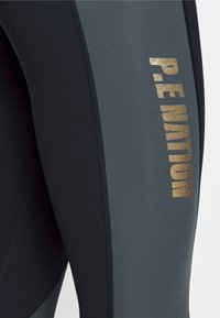 Legginsy z wysokim stanem w czerni i ciemnoszarości, z błyszczącym złotym logo "P.E NATION" z boku oraz płaskimi szwami dla komfortu.