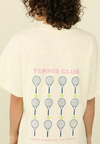 Světle krémové oversized tričko s růžovým textem "TENNIS CLUB" a vzorem modrých tenisových raket a zelených tenisových míčků na zadní straně.