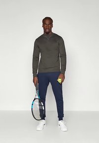 Abbigliamento sportivo con felpa verde scuro e pantaloni blu navy, che tiene in mano una racchetta da tennis e una palla da tennis giallo neon.