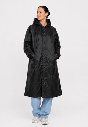Kvinde står med hænderne i lommerne iført en lang sort hættejakke, blå jeans og hvide sneakers mod en ensfarvet baggrund.